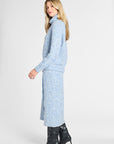 A woman modeling a light blue knit cardigan and matching midi skirt set with a subtle speckled pattern, paired with black heeled ankle boots, against a plain light gray studio background.