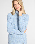 A woman modeling a light blue and white speckled knit mock-neck sweater against a plain light gray studio background.