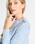 A close-up of a woman wearing a light blue heathered knit sweater with small metallic buttons on the shoulder, set against a plain light gray background.