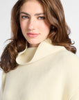 A close-up of a woman with brown hair wearing a cream-colored knit turtleneck sweater against a light gray background.