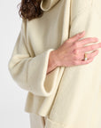 Close-up of a person wearing a cream-colored ribbed knit sweater with a high folded collar and wide cuffs, paired with matching cream pants against a light gray background.