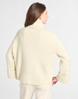 A woman shown from the back wearing a cream-colored knit turtleneck sweater and matching cream trousers against a light gray studio background.