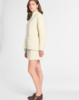A woman modeling a cream-colored knit sweater and matching mini skirt set, paired with brown leather oxford shoes, against a plain light gray studio background.