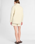 A woman shown from the back wearing a cream-colored knit turtleneck sweater and matching shorts, paired with dark brown leather shoes, against a light gray studio background.