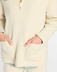 A close-up of a person wearing a cream ribbed knit quarter-zip sweater with front patch pockets, paired with matching knit shorts and a striped shirt peeking out at the hem.