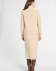 A woman shown from the back wearing a beige knit turtleneck sweater and matching midi skirt, paired with brown knee-high boots, against a light gray studio background.