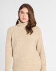 A woman wearing a beige knit turtleneck sweater with raglan sleeves, shown from the waist up against a plain light gray studio background.