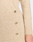 Close-up of a beige knit cardigan or sweater with a fine speckled texture and a vertical row of shiny gold buttons, shown on a model with a hand visible at the side.
