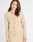 A woman modeling a beige knit cardigan sweater with gold buttons, paired with a matching beige skirt, against a plain light gray studio background.