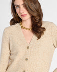 A woman wearing a beige knit cardigan sweater with gold buttons, styled with a chunky gold chain necklace against a light gray background.
