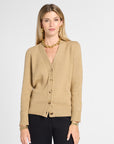 A woman modeling a beige knit cardigan with gold buttons, paired with black pants and gold jewelry, against a light gray studio background.