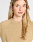 A close-up of a woman with blonde hair wearing a beige knit crewneck sweater and gold hoop earrings against a light gray background.