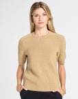 A woman modeling a short-sleeve knit sweater in a warm beige color, paired with black pants, against a light gray studio background.