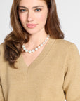 A woman with long brown hair wearing a cream-colored pearl necklace and a beige V-neck sweater, photographed against a light gray studio background.