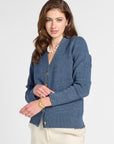 A woman modeling a blue knit cardigan sweater with gold buttons, paired with cream pants and a pearl necklace, against a light gray studio background.