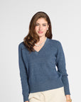 A woman modeling a heathered slate blue V-neck knit sweater with long sleeves, paired with cream pants and a pearl necklace, against a light gray studio background.