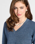 A woman with brown hair wearing a blue V-neck sweater, a white pearl necklace, and a matching white pearl stud earring, photographed against a light gray studio background.