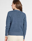 A woman shown from the back wearing a heathered blue knit sweater and cream-colored trousers, photographed against a plain light gray studio background.