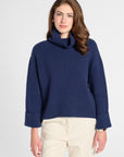 A woman wearing a navy blue knit turtleneck sweater with rolled cuffs and cream-colored trousers, standing against a light gray studio background.