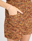 Close-up of a person wearing a brown tweed mini skirt with multicolored yarns, featuring a side pocket and a silver-tone button, paired with a silver bracelet.