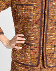 Close-up of a tweed jacket or dress in a multicolor woven blend of brown, gold, red, and cream, with dark brown trim and a hand resting on the hip.