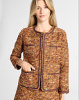 A woman modeling a matching tweed jacket and shorts set in a warm brown, orange, and gold bouclé pattern with dark brown trim, against a light gray studio background.