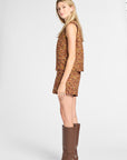 A woman modeling a sleeveless cropped vest and matching shorts set in a textured orange-brown tweed fabric, paired with brown knee-high leather boots, against a light gray studio background.