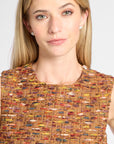 A close-up of a woman wearing a sleeveless textured tweed top with a multicolor pattern of orange, yellow, red, and blue threads, paired with small silver stud earrings, against a light gray background.