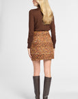 A woman shown from the back wearing a brown long-sleeve top, a high-waisted tweed mini skirt in orange, yellow, and brown tones, and dark brown knee-high boots, against a light gray studio background.