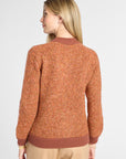 A woman shown from the back wearing a textured, heathered rust-orange knit sweater with solid ribbed trim, paired with beige pants, against a light gray studio background.