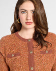 A woman wearing a textured knit cardigan sweater in a warm rust-orange color with brown ribbed trim and gold-tone buttons, shown against a light gray studio background.