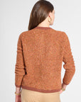 A woman shown from the back wearing a textured, heathered rust-orange knit sweater with brown ribbed trim, paired with beige trousers, against a light gray studio background.