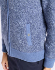 Close-up of a blue heathered knit zip-up cardigan with a ribbed hem and cuffs, featuring a vertical chest pocket with a metal snap button, worn with dark navy pants.