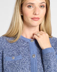 A close-up of a woman wearing a textured blue knit cardigan sweater with silver-tone buttons, set against a plain light gray background.