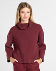 A woman modeling a maroon knit turtleneck sweater and matching trousers against a light gray studio background.