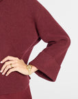 A close-up of a person wearing a ribbed knit long-sleeve top or dress in deep burgundy, with one hand resting on the hip. The person is also wearing a gold watch and ring, set against a clean light gray background.