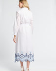Woman modeling a white dress with blue embroidery on the trim on a plain background