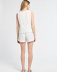 Woman wearing a lace detailed white sleeveless blouse and shorts on a plain background