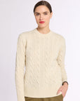 Woman wearing a cream cashmere cable knit sweater and olive green pants on a white background with hand in pocket