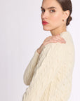 Portrait of woman with hand on shoulder wearing a cashmere cream cable knit crew sweater on a white background