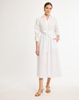 A woman standing in a studio setting, wearing a white shirt dress with a sash tied at the waist, made of Italian fabrics.