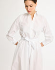 A medium shot of a woman looking down, wearing a white, long-sleeve shirt dress made of Italian fabrics, with a matching sash tied at the waist. Her hands are resting in the dress pockets.