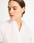 A close-up portrait of the model's head and shoulders wearing the white, collared Eleanor Shirt Dress. She has her hair pulled back and is looking down and to the side, highlighting the crisp button-front placket and neckline.