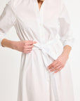 A woman standing, tying the sash of a white, long-sleeve shirt dress, made of Italian fabrics.