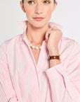 Woman wearing a pink striped shirt with a pearl necklace and brown bracelet on a light gray background