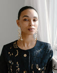 A close-up of a woman's profile wearing large, gold-toned, floral-shaped chandelier earrings. She is wearing a dark, patterned dress.