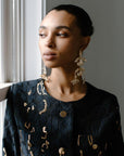 A close-up of a woman's profile wearing large, gold-toned, floral-shaped chandelier earrings. She is wearing a dark, patterned dress and is looking out a window.