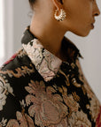 A close-up profile view of a woman's face and shoulder wearing gold scalloped hoop earrings. She is wearing a dark floral or paisley patterned blouse.