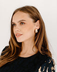 A medium close-up of a woman's face and shoulder wearing thick, gold oval hoop earrings. She is wearing a dark, patterned top and looking up and to the left.
