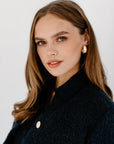 A medium close-up of a woman's face and shoulder wearing thick, gold oval hoop earrings. She is wearing a dark, patterned top and looking up and to the left.